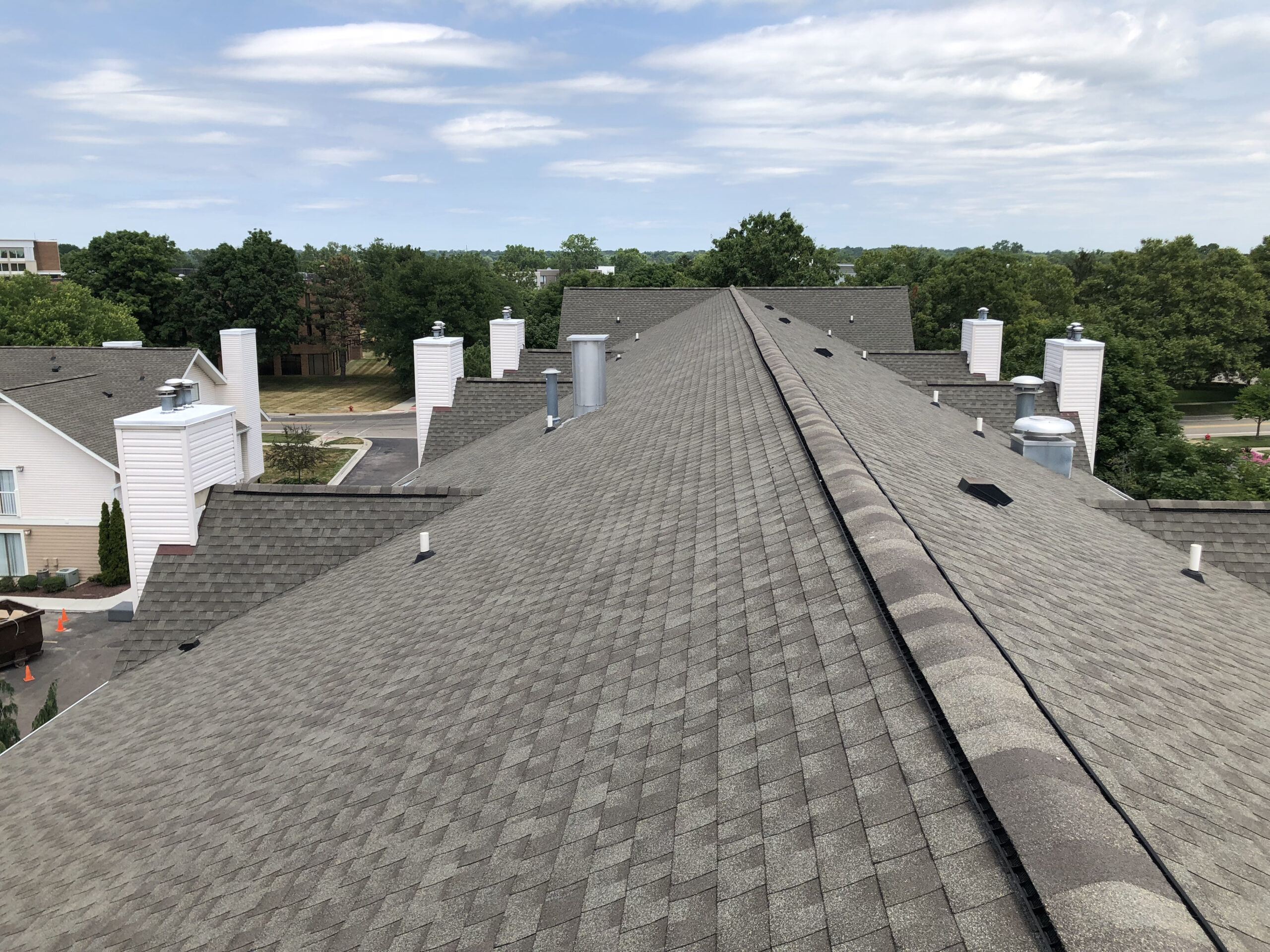 Aerial view of a gray shingled roof with multiple chimneys and a row of townhouses surrounded by trees under a cloudy sky.