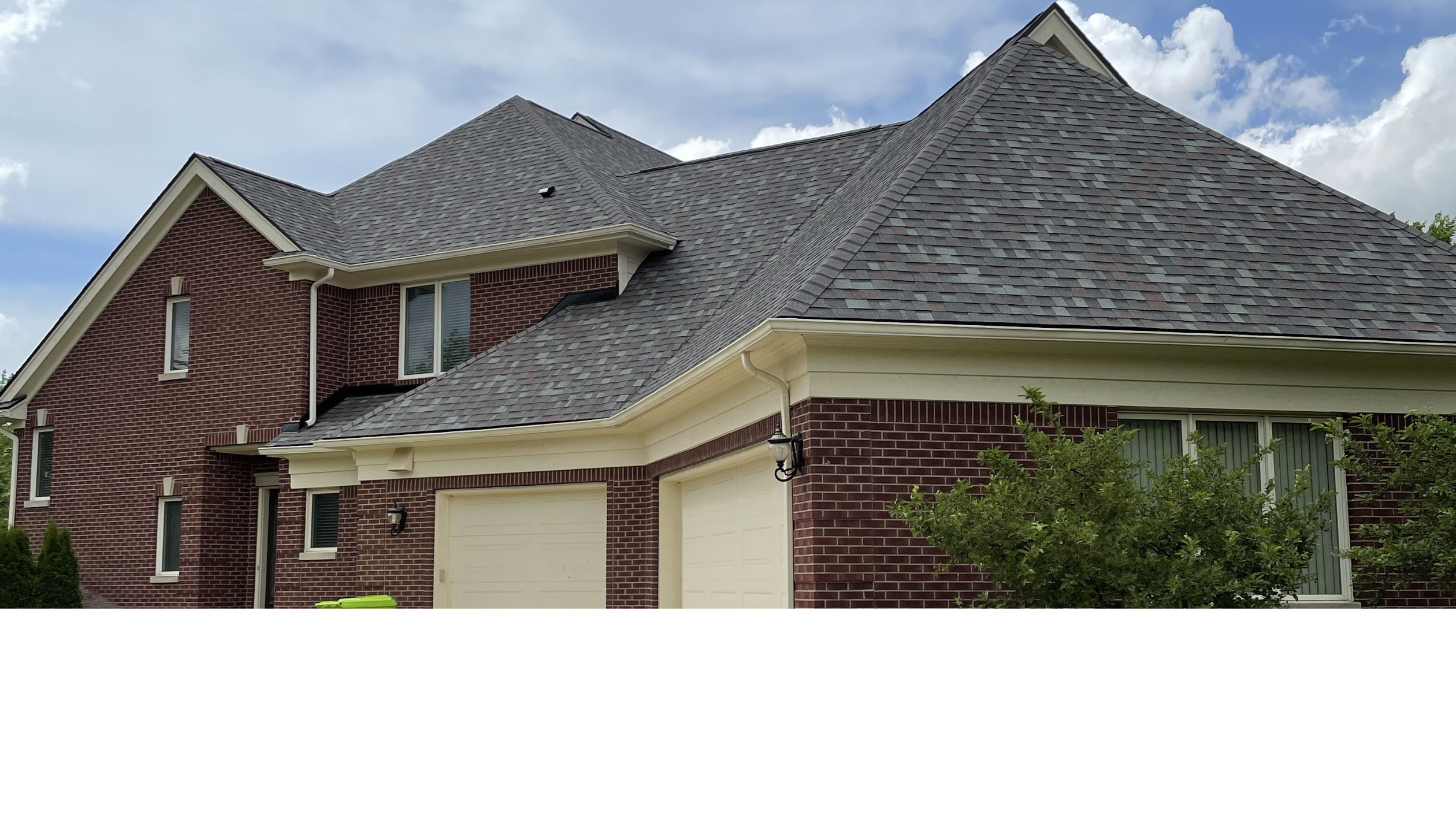 A large brick house with a steep, shingled roof, featuring multiple gables and a double garage under a cloudy sky.