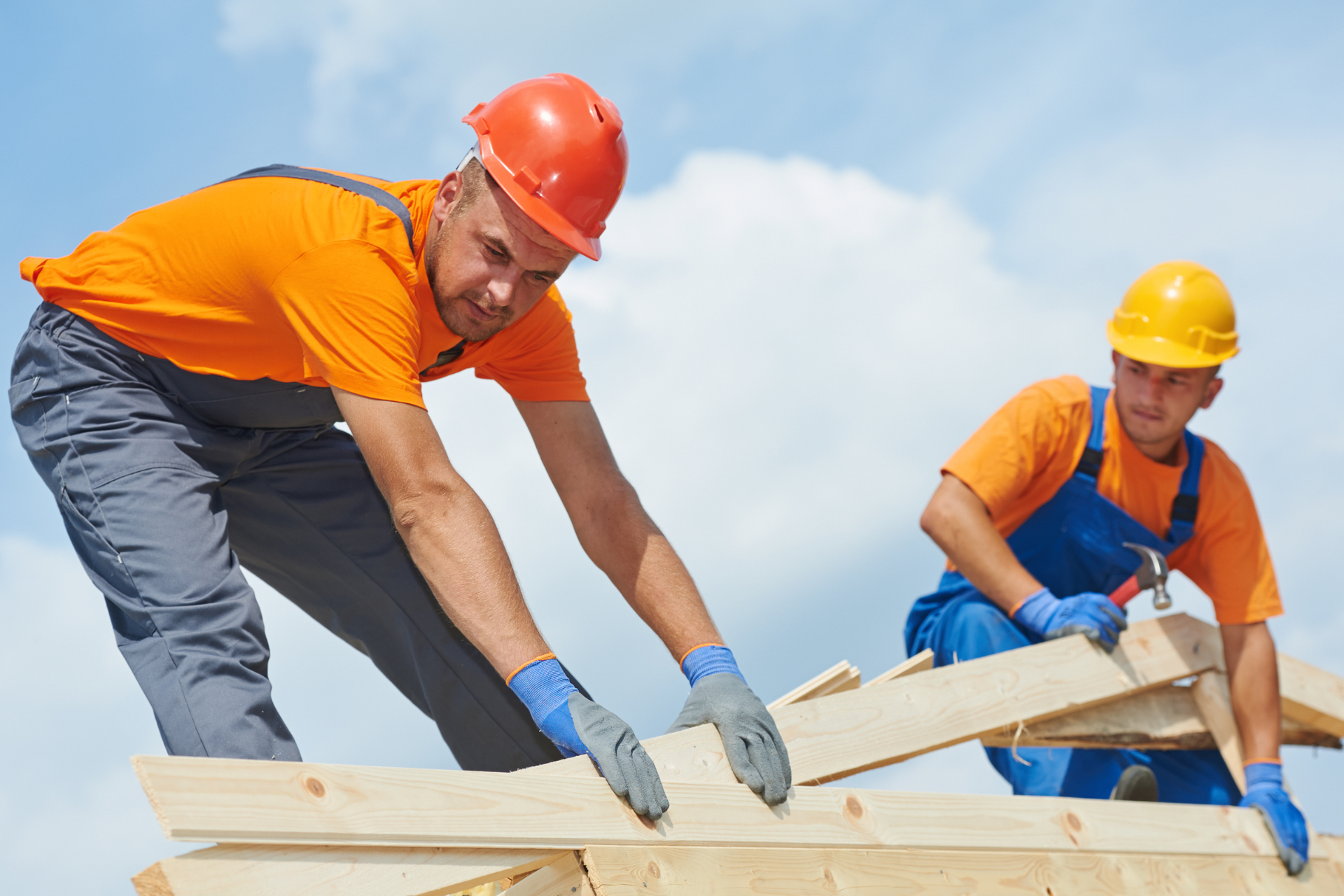 Two construction workers in orange shirts and hard hats assembling wooden beams on a clear day.