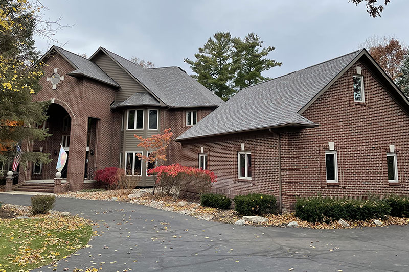 Large brick house with multiple roofs, including a prominent arched entryway, surrounded by autumn foliage and a paved driveway.