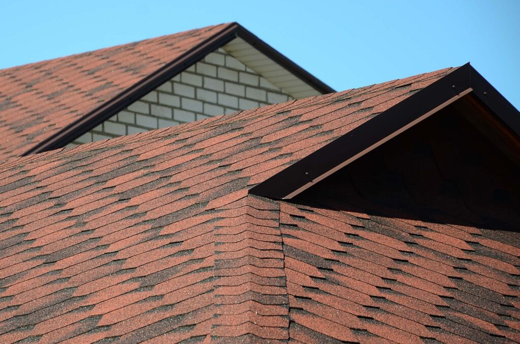 Close-up view of a house's roof with brown asphalt shingles and a small dormer against a clear blue sky.