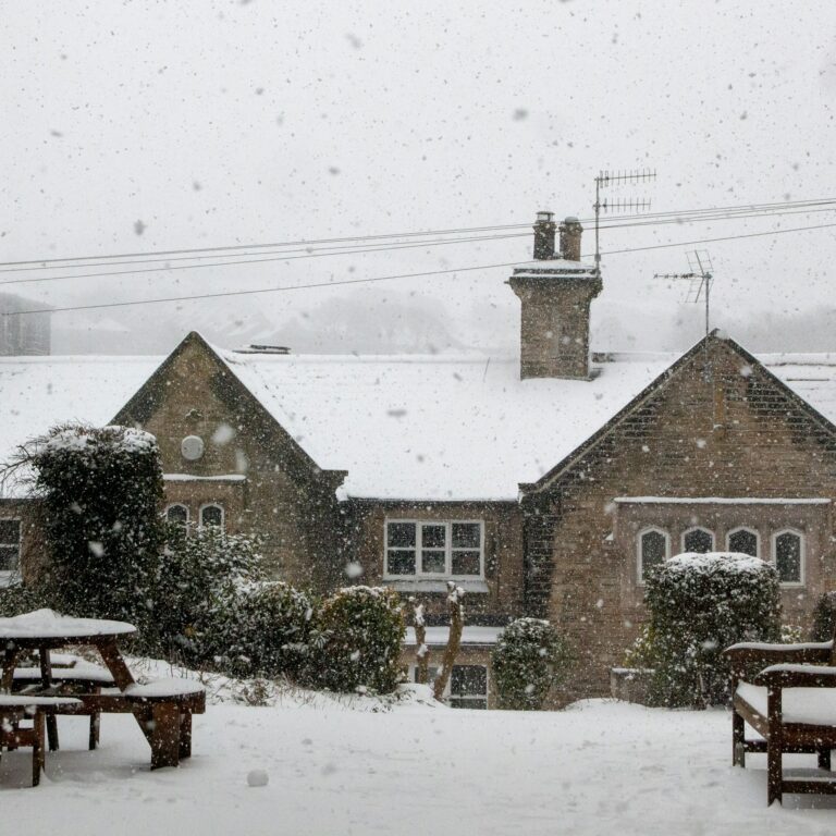Heavy snowfall over a rural stone house with snow-covered benches and a garden, visibly accumulating snowflakes in the air.