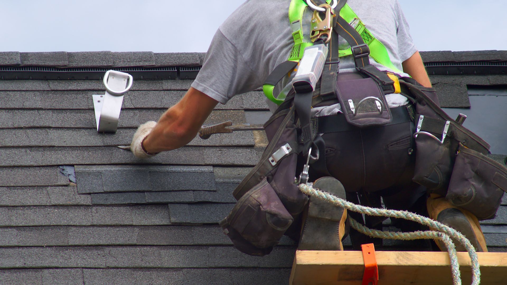 A worker wearing safety gear installs shingles on a roof, using tools from a belt.