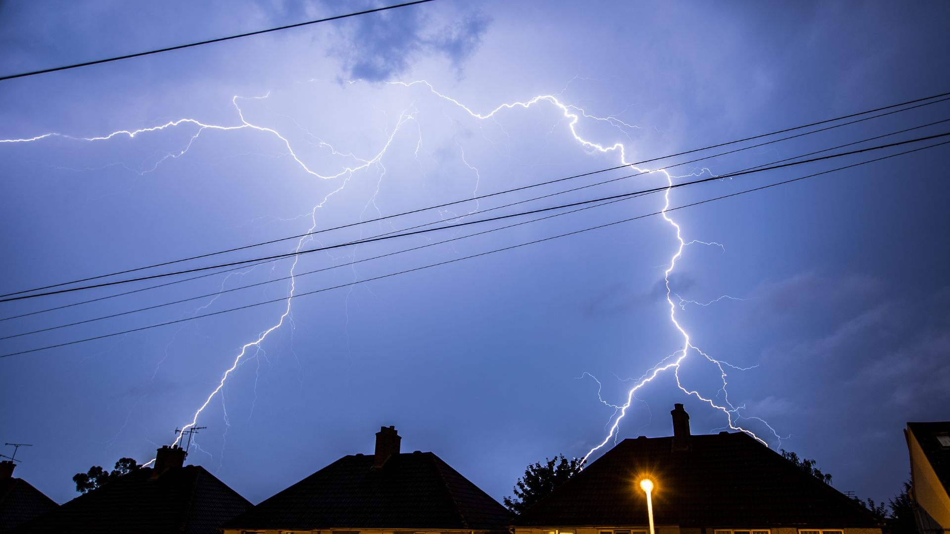 Lightning strikes in the night sky above houses with silhouetted rooftops and power lines.