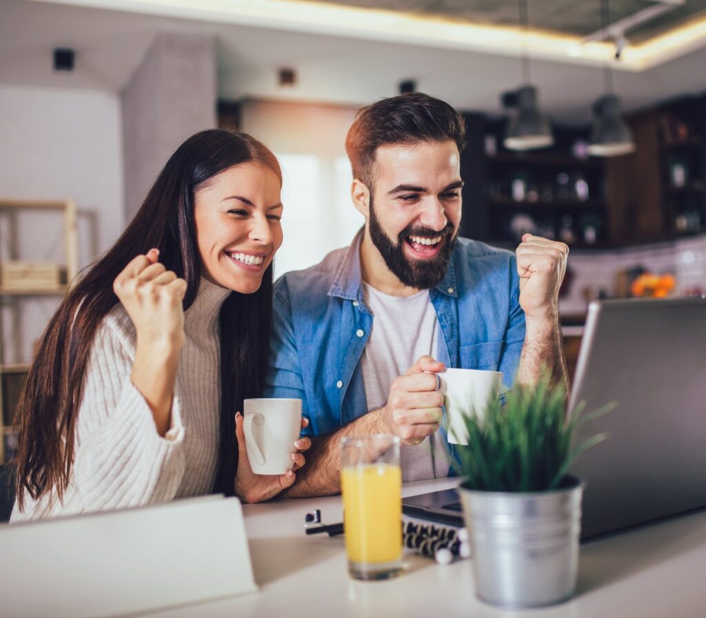 A man and woman smile and raise their fists in excitement while looking at a laptop on a table with a plant, glasses, and a glass of juice.