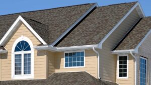 asphalt shingle roofing in michigan The image shows the exterior of a house with beige siding, white trim, large windows, and a dark shingle roof under a clear blue sky.