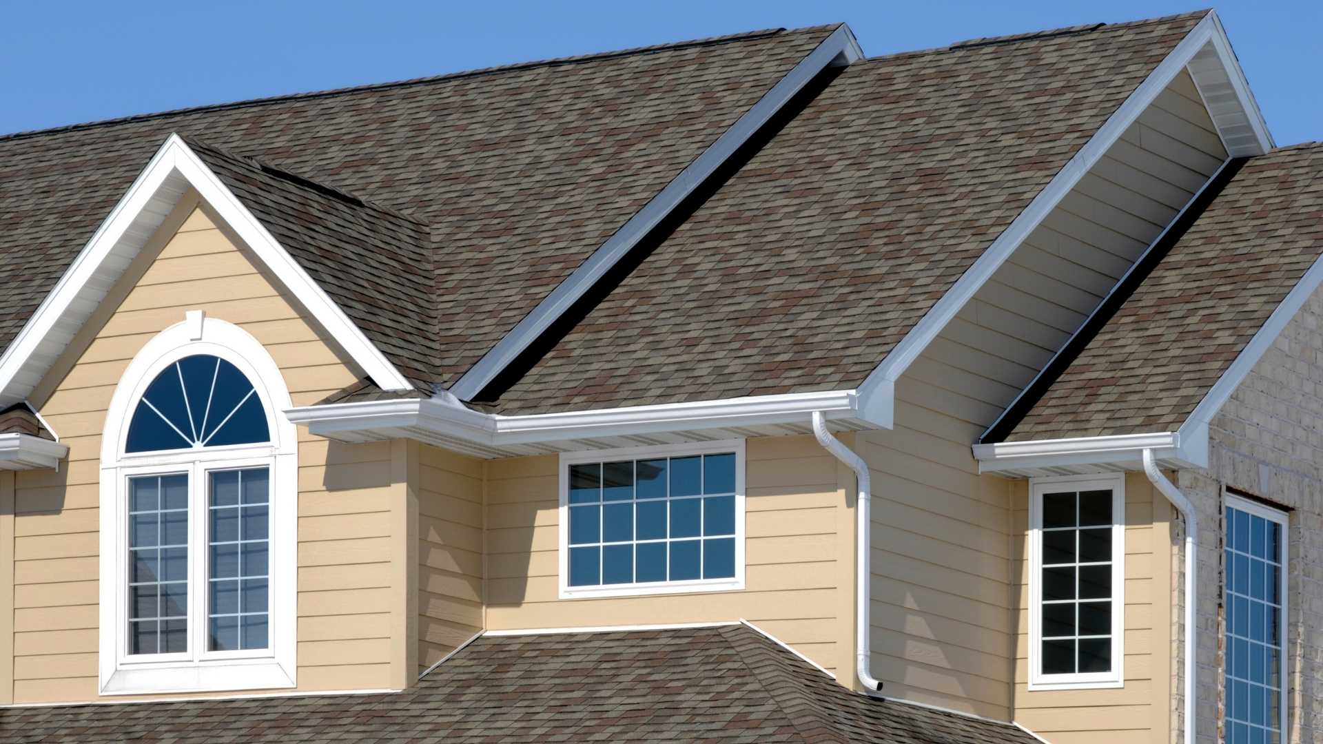 asphalt shingle roofing in michigan The image shows the exterior of a house with beige siding, white trim, large windows, and a dark shingle roof under a clear blue sky.