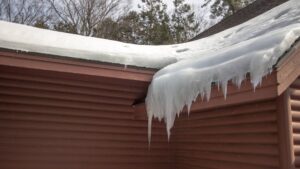 Prevent ice dams in michigan Large icicles and a thick layer of snow hang from the edge of a brown house roof on a cold winter day.