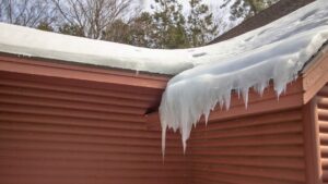 ice dam prevention in michigan Large icicles hang from the edge of a snow-covered roof on a red wooden house, with bare trees and evergreens visible in the background.