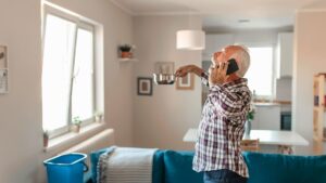 roof leak repair in michigan Older man standing in a living room, talking on the phone, holding a small pot, with a blue bucket on the floor near a window.