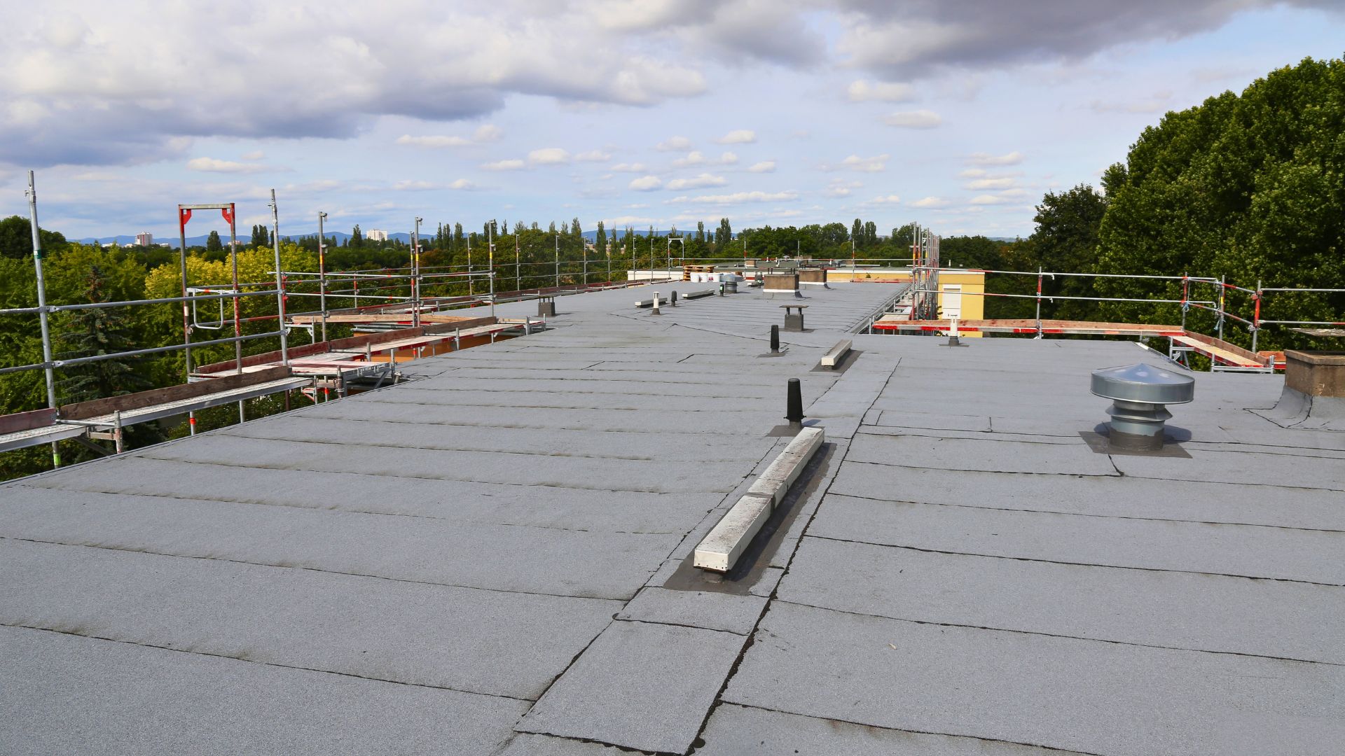 Michigan comemercial roofing Flat rooftop with grey surface, surrounded by safety railings, ventilation pipes, and a metal vent; trees and clouds visible in the background.